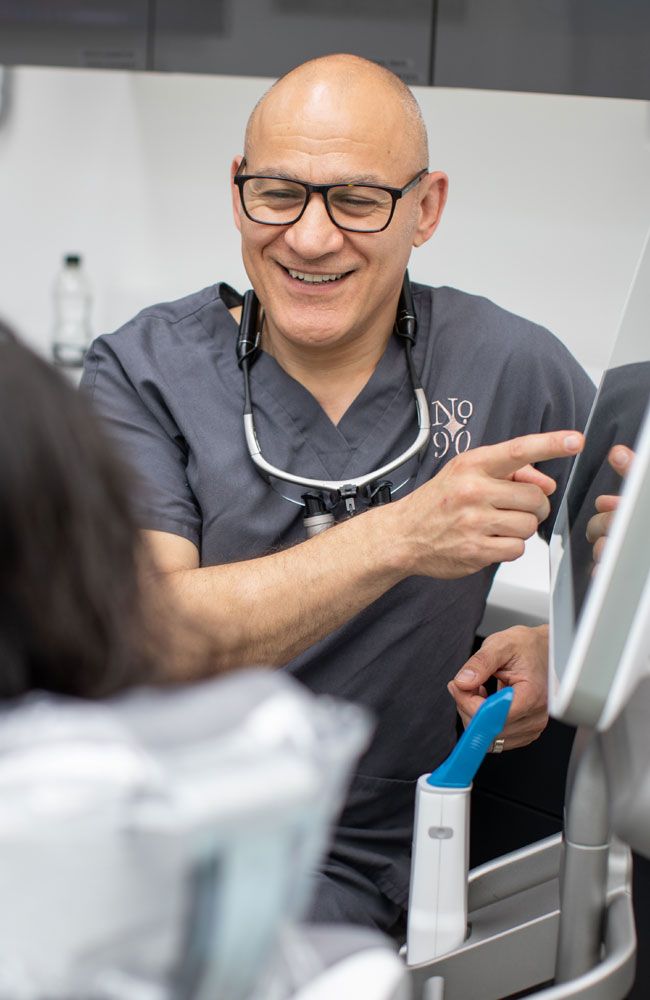A smiling male dentist in grey scrubs and loupes points to a screen.