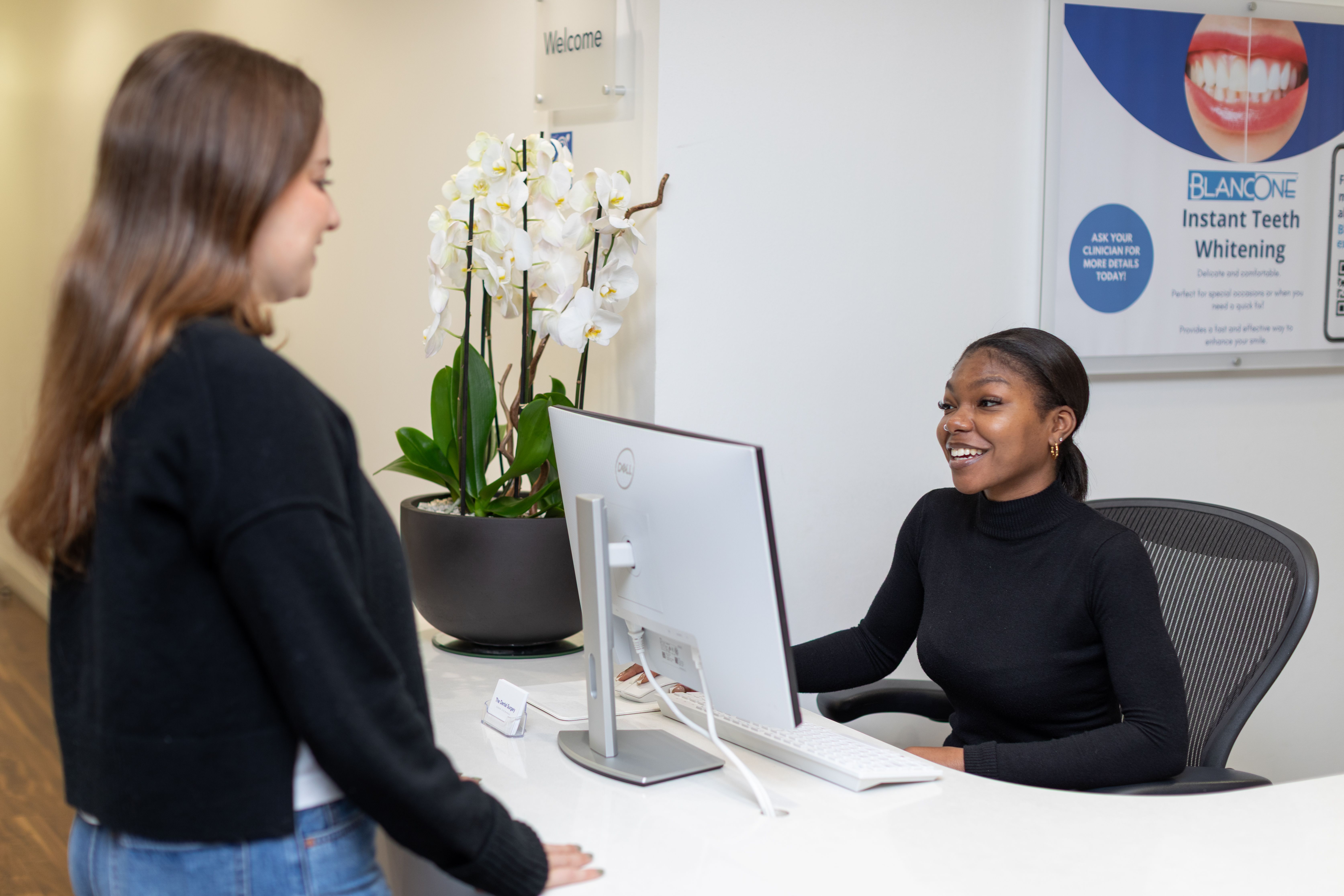 A smiling receptionist at a desk speaks with a patient in a dental office.