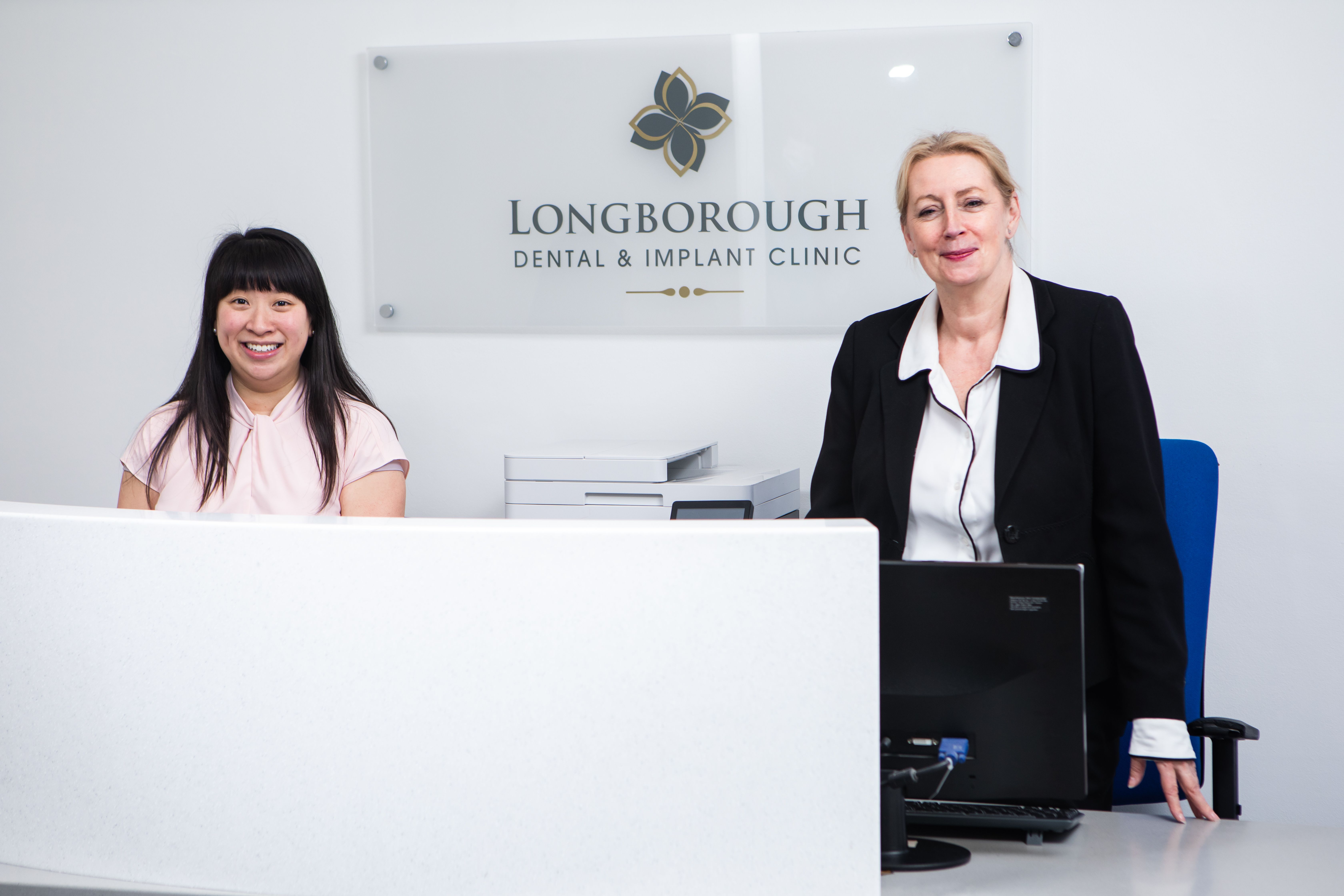two women are standing at a longborough dental and implant clinic reception desk .
