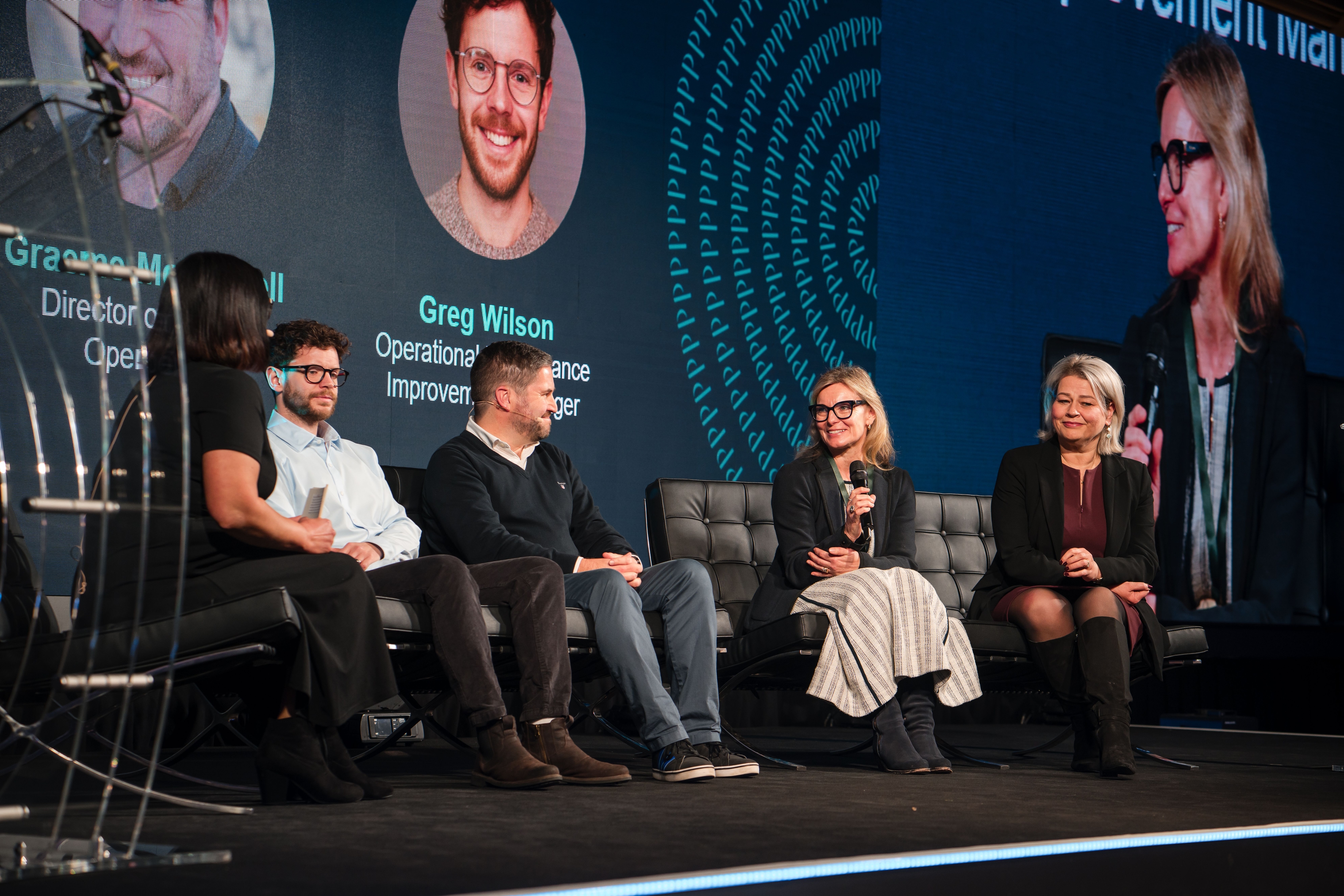 Five panelists on a stage with a large screen behind them displaying presenter information.